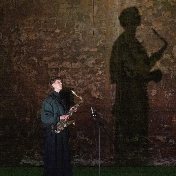Heli Hartikainen esiintyy tenorisaksofonilla, varjo heijastuu tiiliseinään Chronovariations-esityksessä Kiisuhallilla